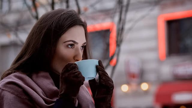 Mulher tomando café em uma xícara na rua, num dia frio, com luzes desfocadas ao fundo