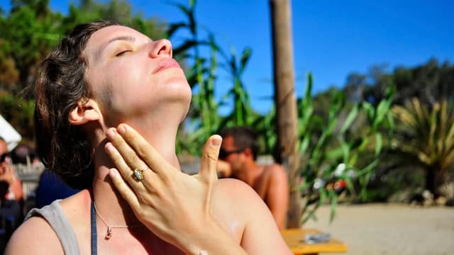 Mulher de olhos fechados passando protetor no pescoço, com o sol forte no rosto