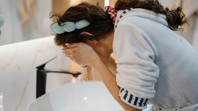 Mulher de roupão e faixa no cabelo lavando o rosto com as mãos na pia do banheiro