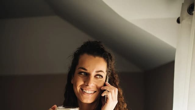 Mulher sorrindo com uma caneca de café na mão ao lado da janela, em um quarto com luz da manhã