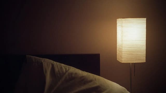 Abajur aceso ao lado da cama em um quarto escuro, com luz quente e baixa