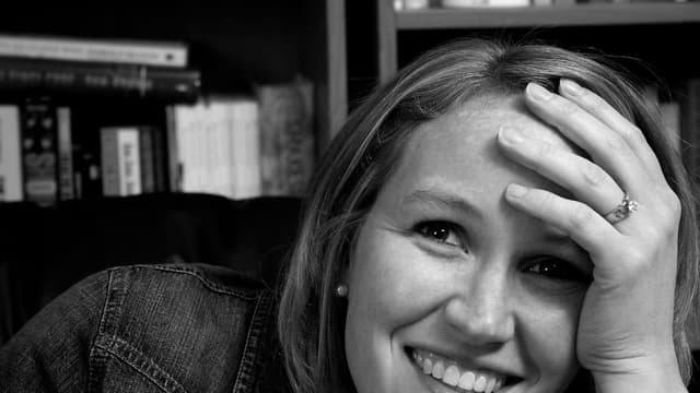 Mulher sorrindo com a mão na testa, em preto e branco, diante de uma estante de livros