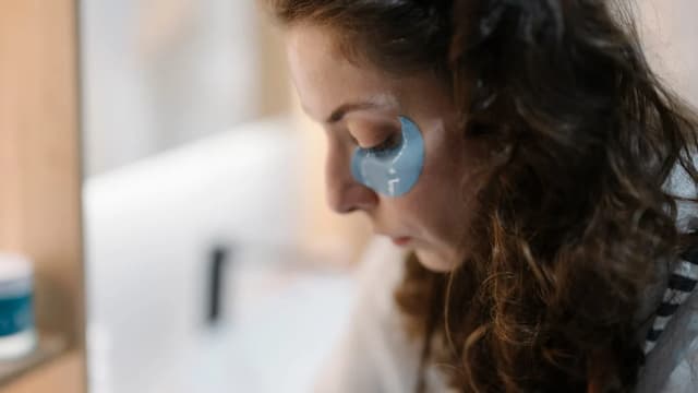 Close do rosto de uma mulher de olhos fechados com adesivos de gel sob os olhos, no banheiro