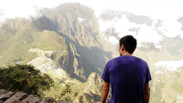 Homem de camiseta roxa sentado de costas em uma pedra, olhando as montanhas e as ruínas lá embaixo