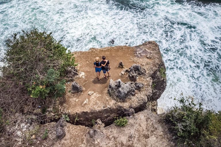 Vista aérea de um casal em pé sobre um penhasco com o mar batendo embaixo