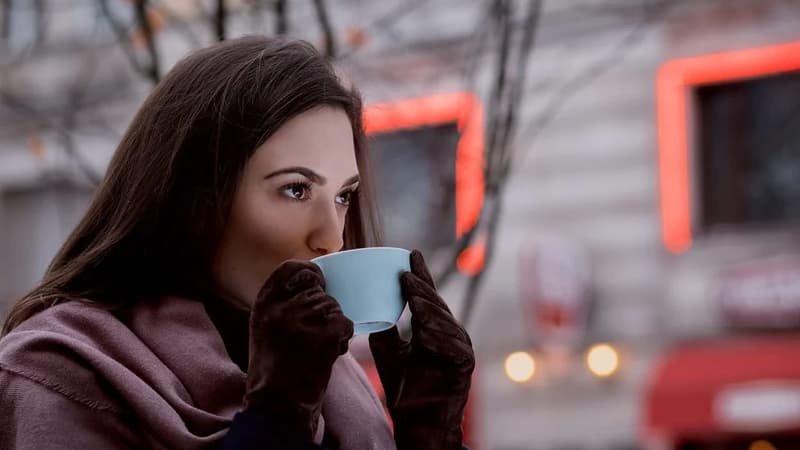 Mulher tomando café em uma xícara na rua, num dia frio, com luzes desfocadas ao fundo