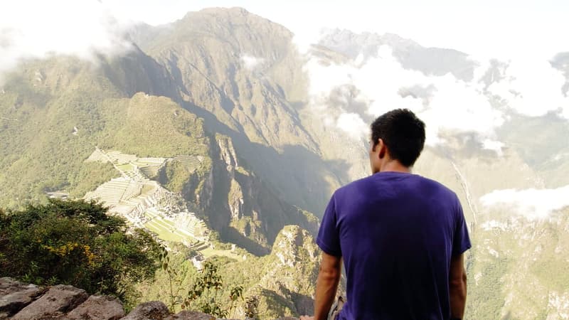 Homem de camiseta roxa sentado de costas em uma pedra, olhando as montanhas e as ruínas lá embaixo